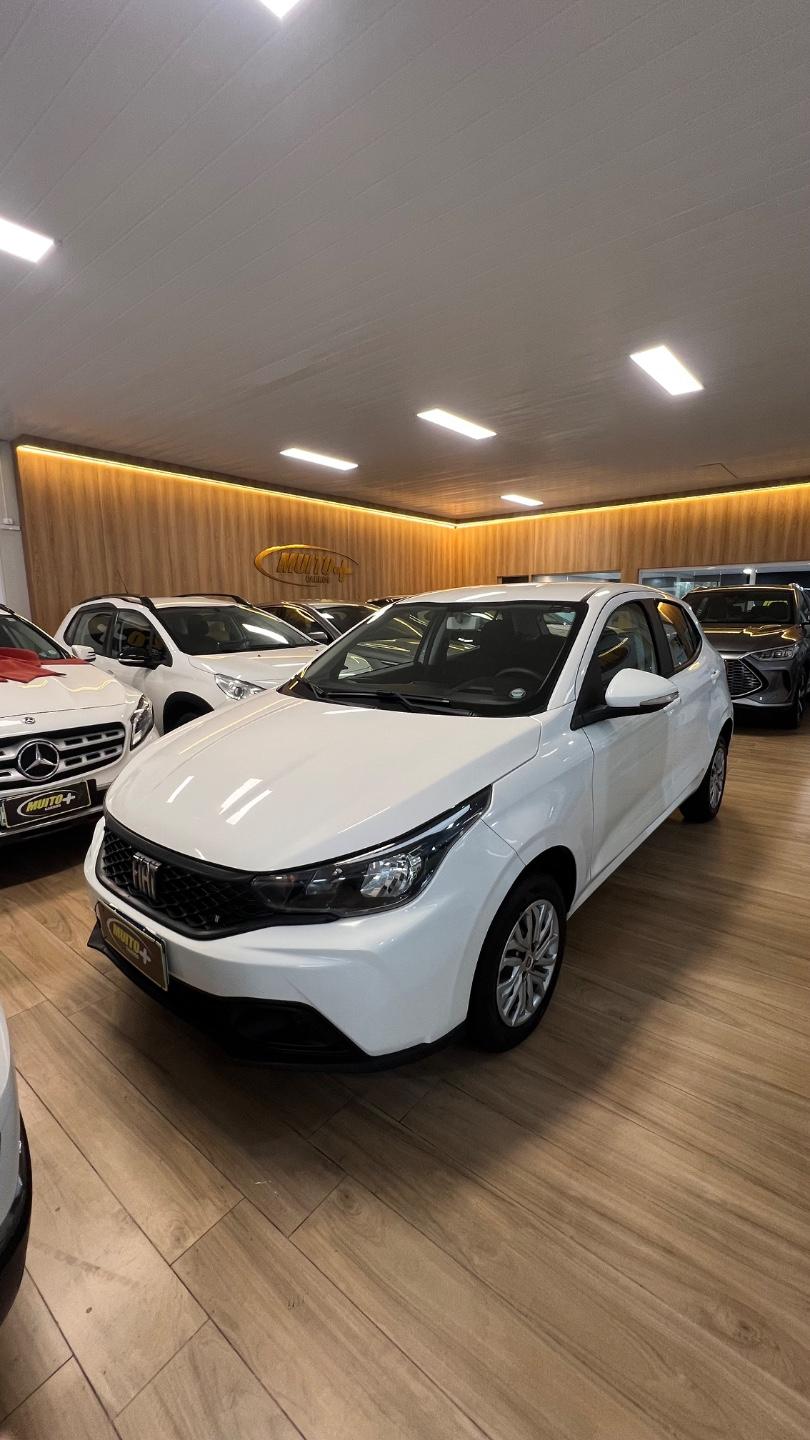 Hatch branco da Fiat visto de frente e de lado no showroom com piso de madeira, com outros carros expostos ao fundo