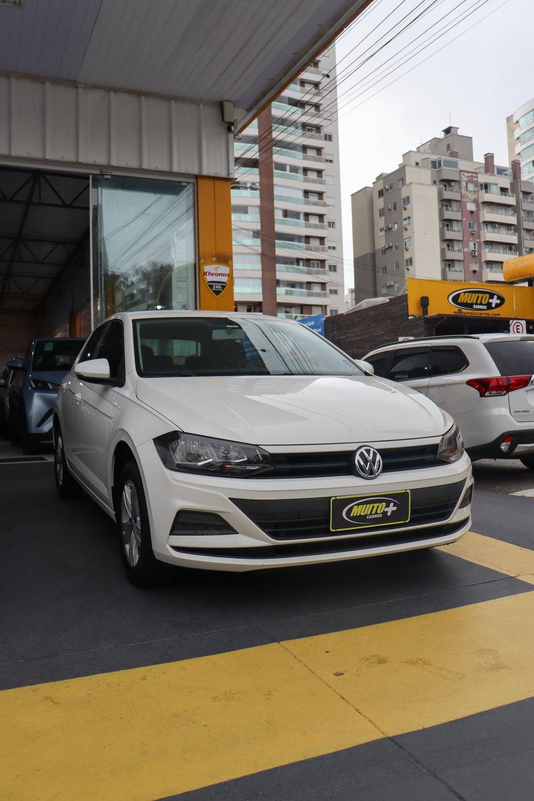 Hatch Volkswagen branco visto de frente no pátio da Muito Mais Carros, com outros veículos e prédios ao fundo
