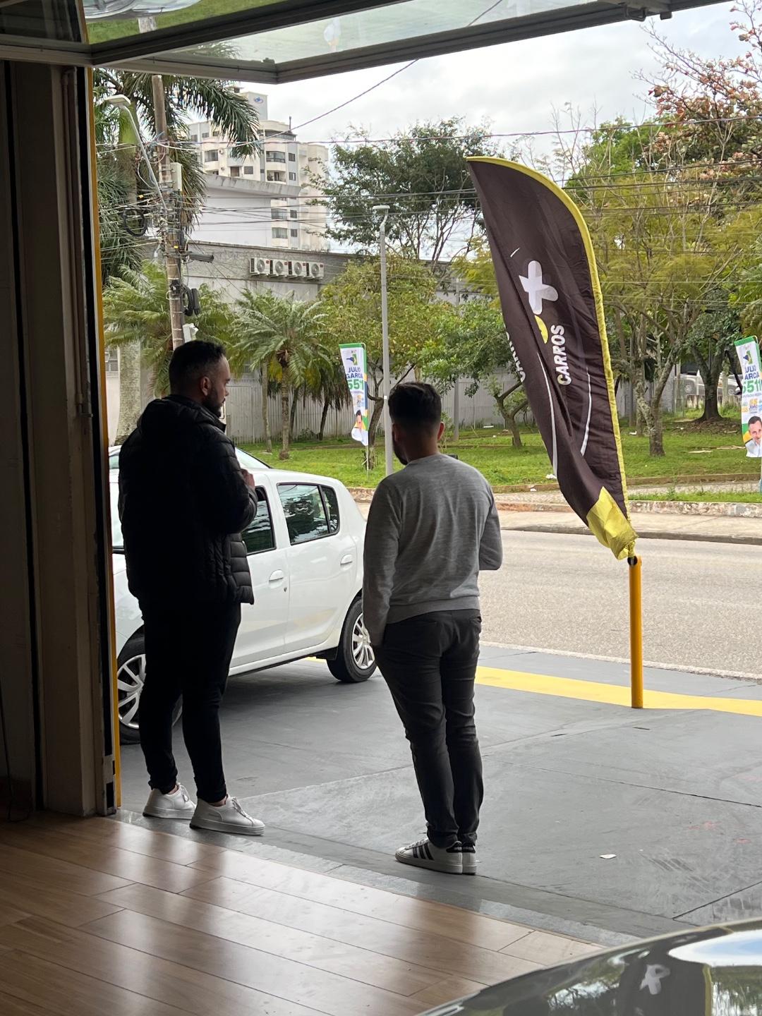 Vista de dentro da loja, pela porta aberta, de dois homens de costas conversando na calçada em frente a um hatch branco estacionado, ao lado de um wind banner com a palavra Carros