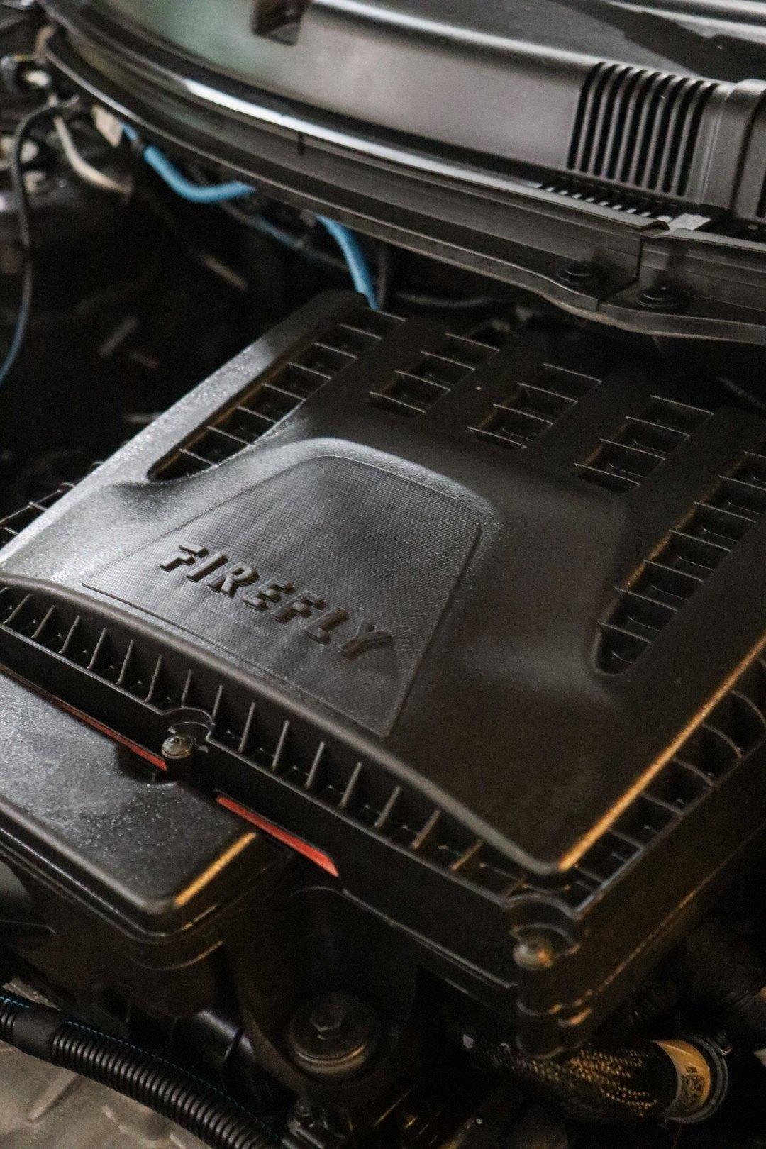 Compartimento do motor com o capô aberto, mostrando a tampa plástica preta com a inscrição FIREFLY, mangueiras e chicotes elétricos