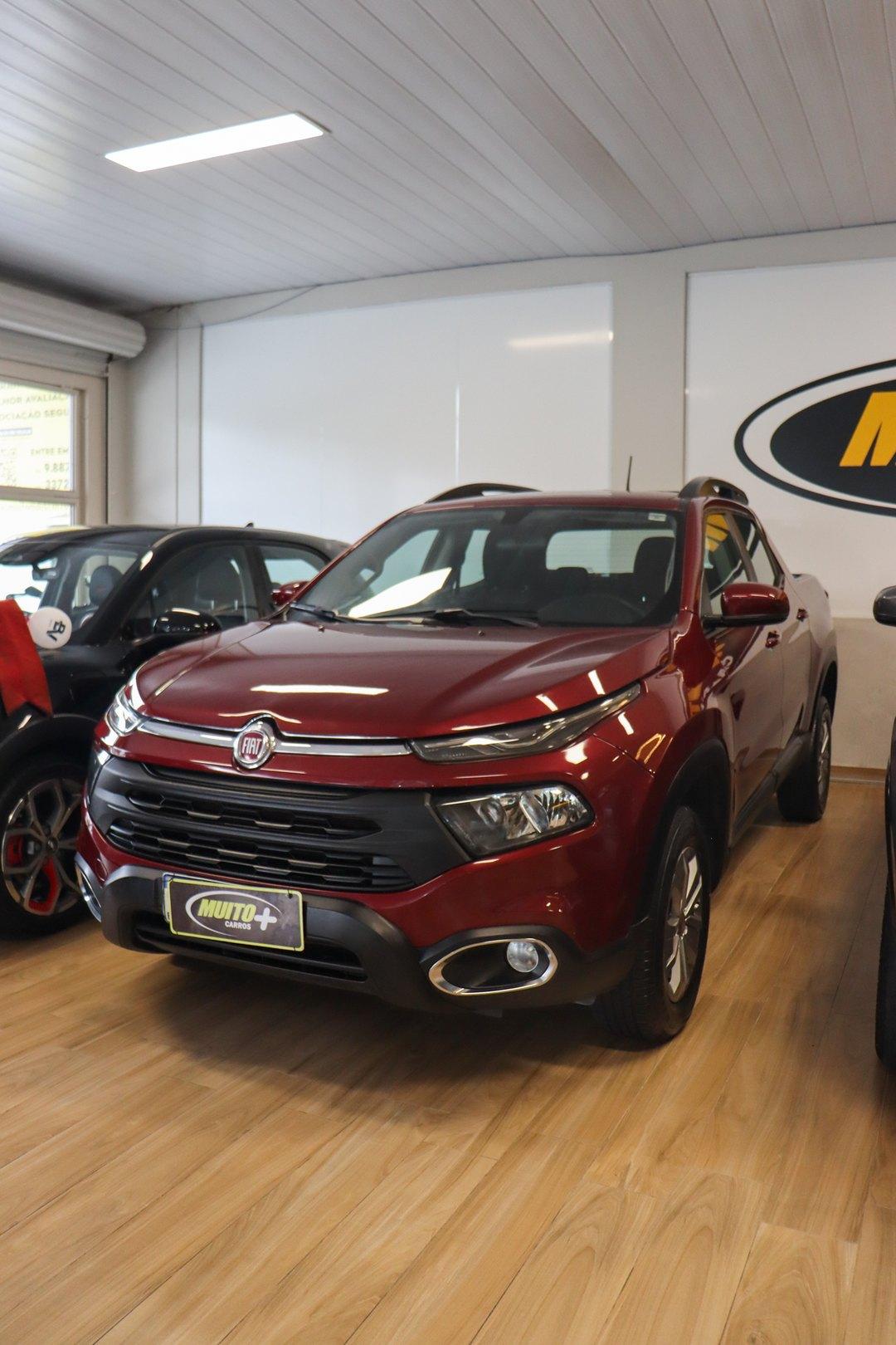 Picape Fiat vermelha de cabine dupla vista de frente em ângulo, exposta no showroom interno da Muito Mais Carros com piso de madeira