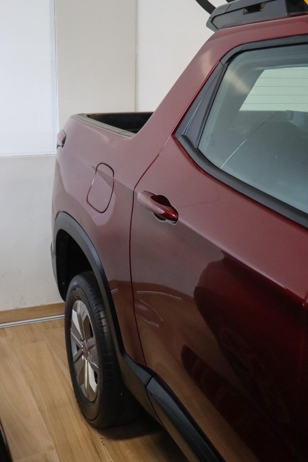 Lateral traseira de uma picape cabine dupla vermelha em ambiente interno com piso de madeira, mostrando a porta traseira, a roda de liga leve e a caçamba